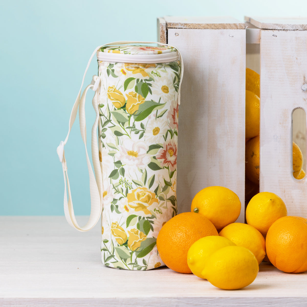 A floral bottle bag with a white strap stands on a white tabletop beside white wooden crates and bright lemons and oranges against a pale blue background.