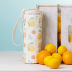 A fruit print bottle bag with a white strap stands on a white tabletop beside white wooden crates and bright lemons and oranges against a pale blue background.