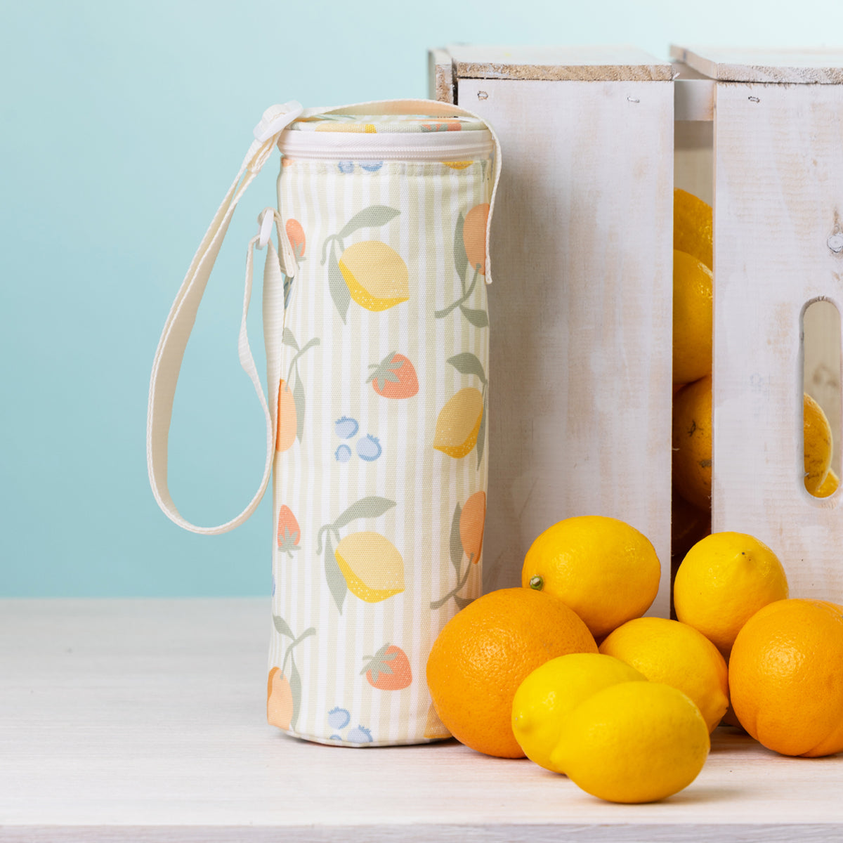 A fruit print bottle bag with a white strap stands on a white tabletop beside white wooden crates and bright lemons and oranges against a pale blue background.