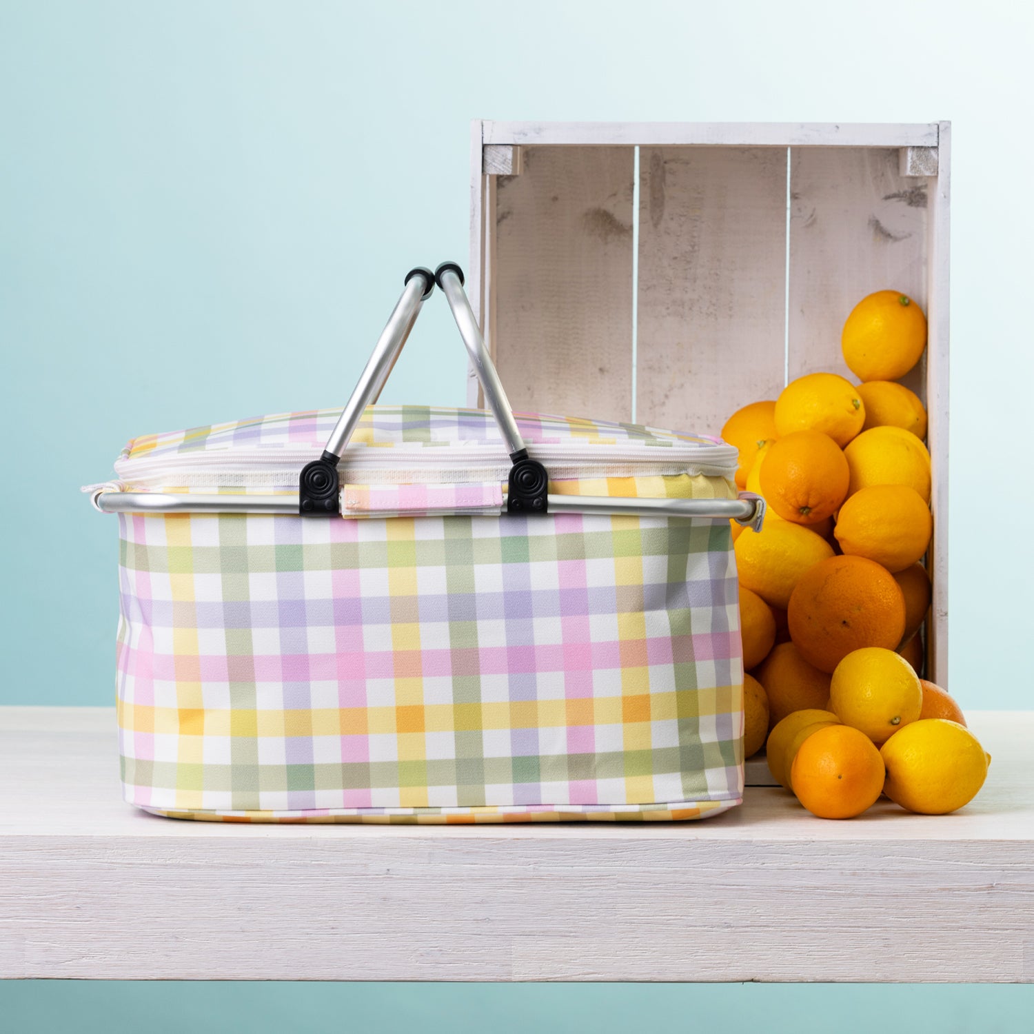 A check picnic cooler basket sits on a white wooden table next to a white crate filled with oranges, against a light blue background.