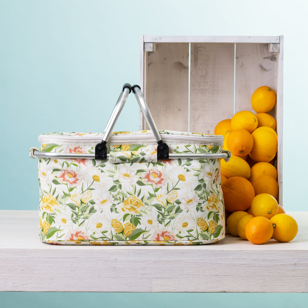 A floral picnic cooler basket sits on a white wooden table next to a white crate filled with oranges, against a light blue background.