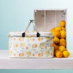 A fruit print picnic cooler basket sits on a white wooden table next to a white crate filled with oranges, against a light blue background.