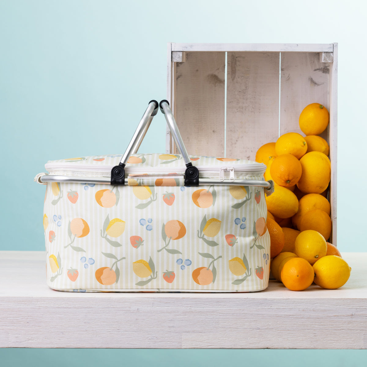 A fruit print picnic cooler basket sits on a white wooden table next to a white crate filled with oranges, against a light blue background.