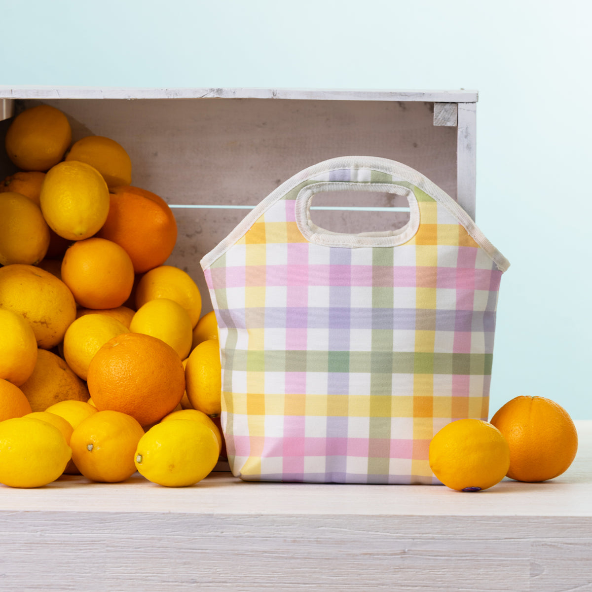 A pastel plaid lunch tote with top handles sits on a light wood surface beside a crate of lemons and oranges against a pale blue background.