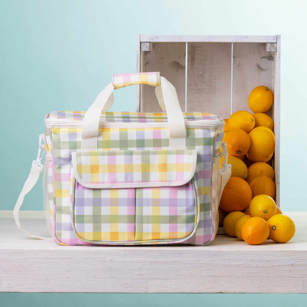 A check cooler bag sits on a white wooden table next to a white crate filled with oranges, against a light blue background.