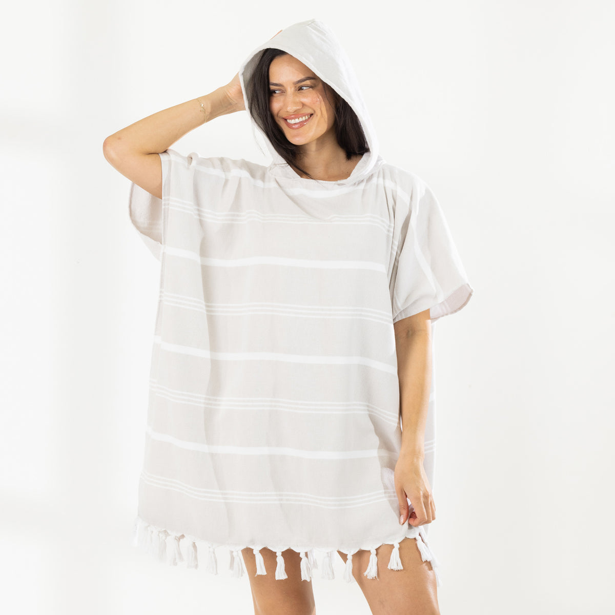 A woman wearing a classic stripe hooded beach poncho with fringed tassels along the hem, photographed against a clean white studio background.