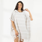 A woman wearing a classic stripe hooded beach poncho with fringed tassels along the hem, photographed against a clean white studio background.