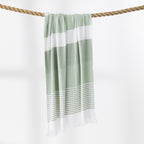 A green and white striped beach towel with fringed edges hangs from a thick rope against a clean white background.