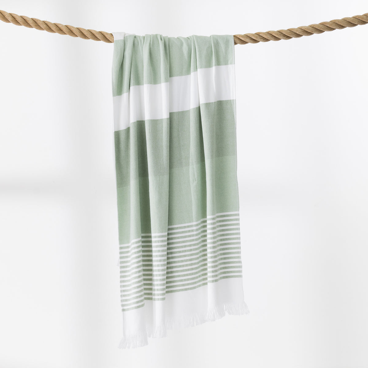 A green and white striped beach towel with fringed edges hangs from a thick rope against a clean white background.