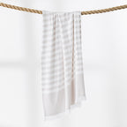 A natural and white striped beach towel with fringed edges hangs from a thick rope against a clean white background.