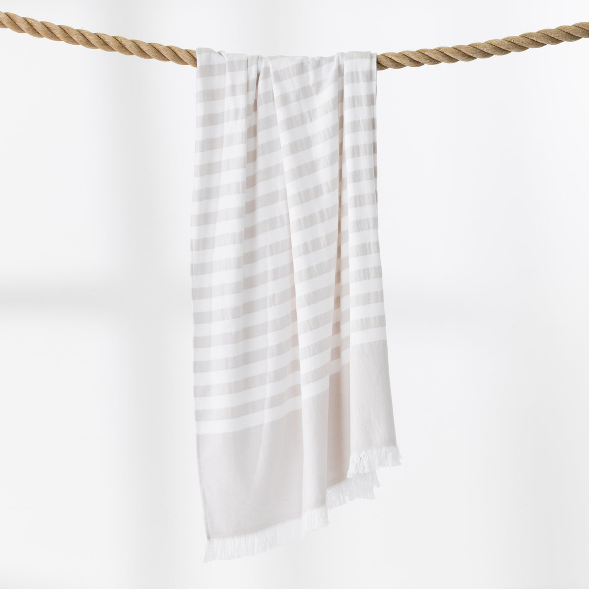A natural and white striped beach towel with fringed edges hangs from a thick rope against a clean white background.
