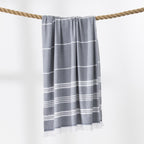 A grey and white striped beach towel with fringed edges hangs from a thick rope against a clean white background.