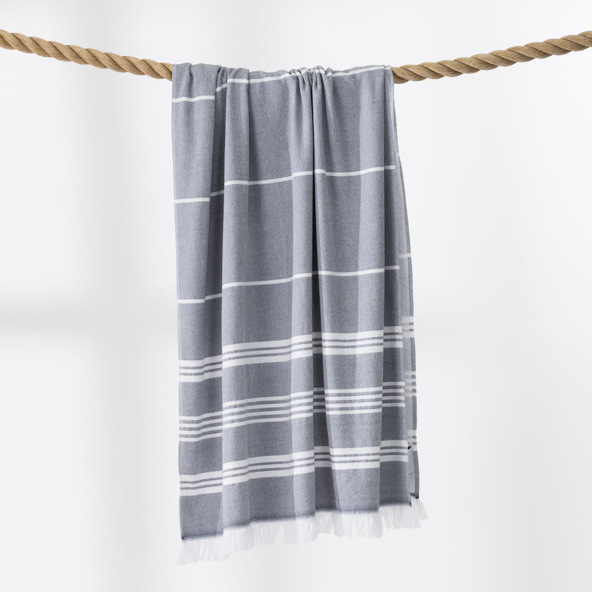 A grey and white striped beach towel with fringed edges hangs from a thick rope against a clean white background.