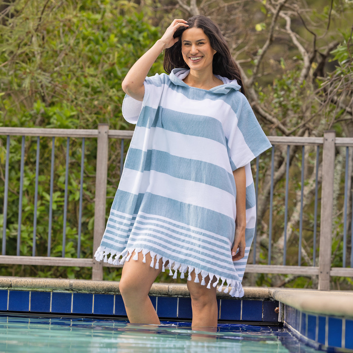 A woman stands in a swimming pool wearing a striped hooded beach poncho with fringe trim, with a wooden fence and green foliage in the background.