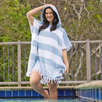 A woman stands in a swimming pool wearing a striped hooded beach poncho with fringe trim, with a wooden fence and green foliage in the background.