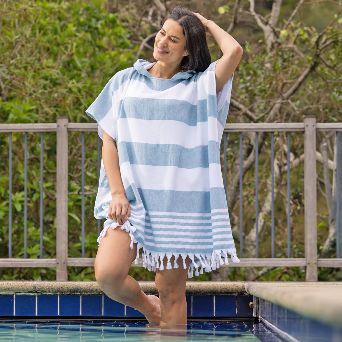 A woman stands in a swimming pool wearing a striped hooded beach poncho with fringe trim, with a wooden fence and green foliage in the background.