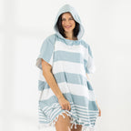 A woman wearing a classic stripe hooded beach poncho with fringed tassels along the hem, photographed against a clean white studio background.