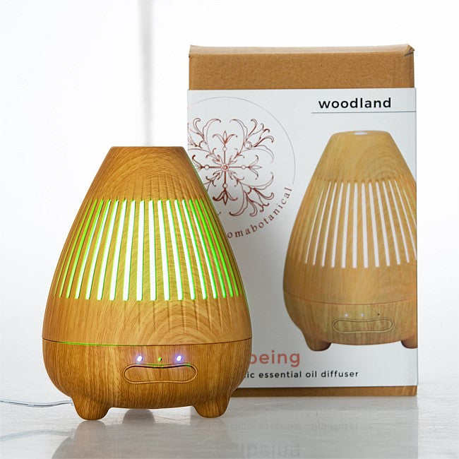 Wooden essential oil diffuser lamp with green light glowing from the inside, shown next to its cardboard packaging box labeled 'woodland'.