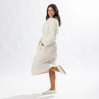 Woman wearing a cream textured plush bathrobe with a shawl collar, self-tie belt, patch pockets and matching cream slippers, posing in a side profile.