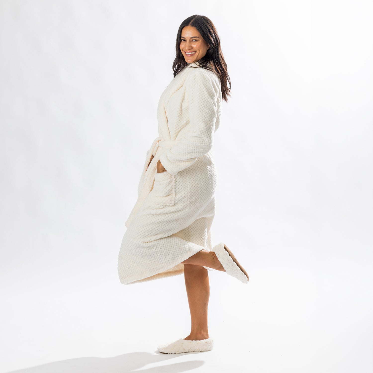 Woman wearing a cream textured plush bathrobe with a shawl collar, self-tie belt, patch pockets and matching cream slippers, posing in a side profile.