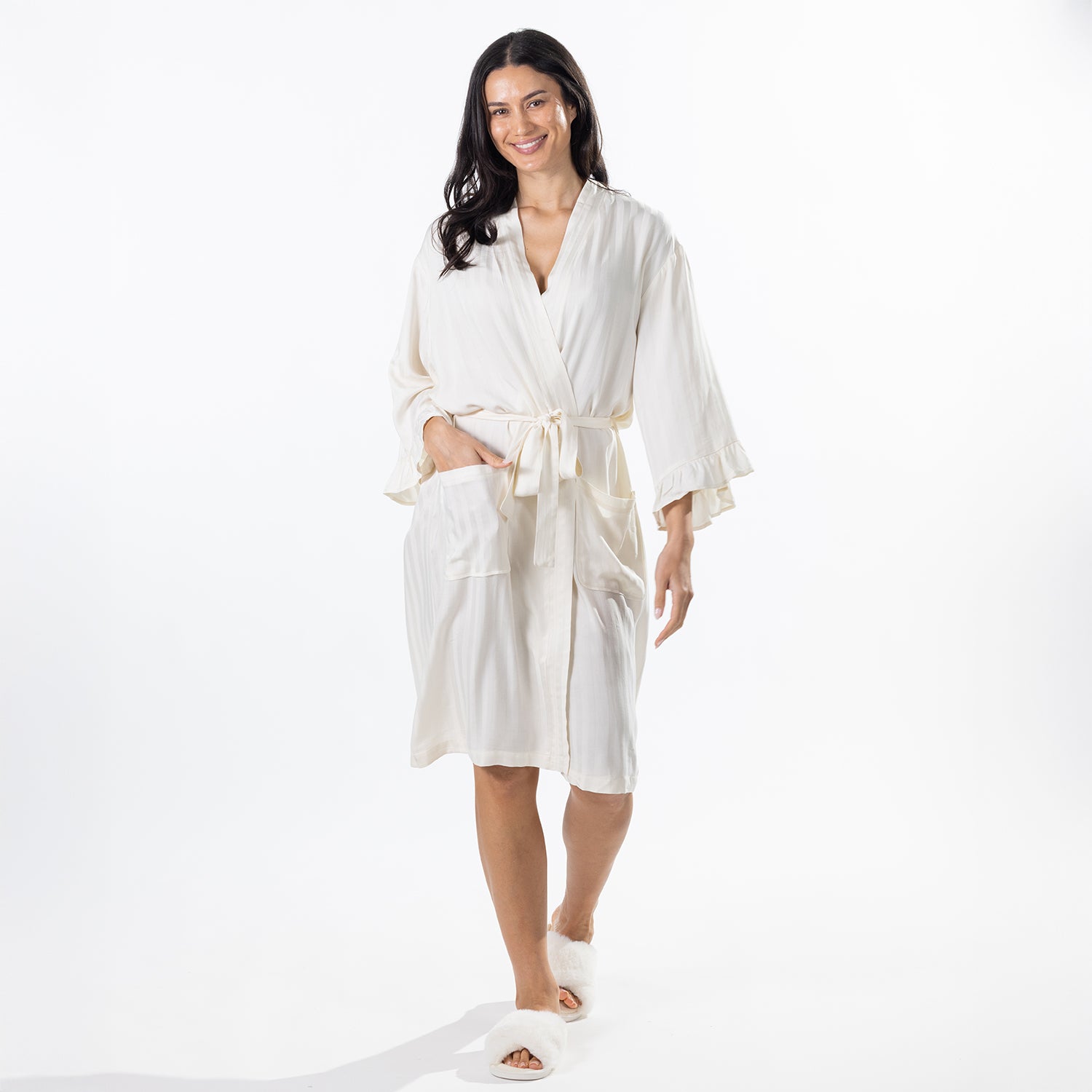 A woman modeling a cream-colored cotton robe with a tie waist and wide sleeves, paired with white slippers, against a solid white studio background.