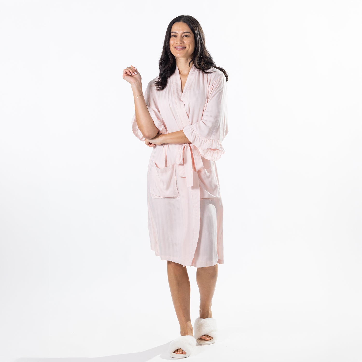 A woman modeling a light pink wrap robe with a tie waist and front pockets, paired with white slippers, against a plain white studio background.
