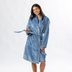 A person wearing a soft, plush pale blue bathrobe with a waist tie is standing and smiling against a plain light background.