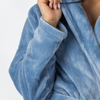 A close-up view of a person wearing a plush pale-blue bathrobe highlights the robe’s soft texture, shawl collar, and sleeve.