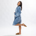 A person wearing a soft, plush pale blue bathrobe with a waist tie is standing and smiling against a plain light background.
