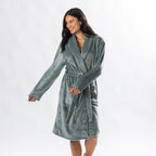 A person wearing a soft, plush sage bathrobe with a waist tie is standing and smiling against a plain light background.