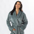 A person wearing a soft, plush sage bathrobe with a waist tie is standing and smiling against a plain light background.