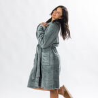 A person wearing a soft, plush sage bathrobe with a waist tie is standing and smiling against a plain light background.