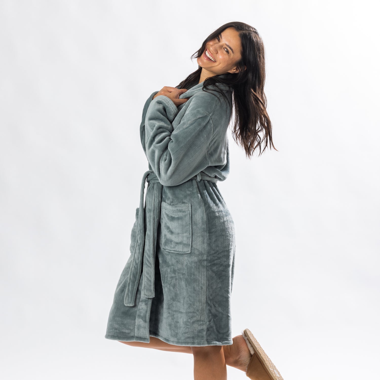 A person wearing a soft, plush sage bathrobe with a waist tie is standing and smiling against a plain light background.