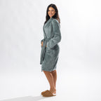 A person wearing a soft, plush sage bathrobe with a waist tie is standing and smiling against a plain light background.