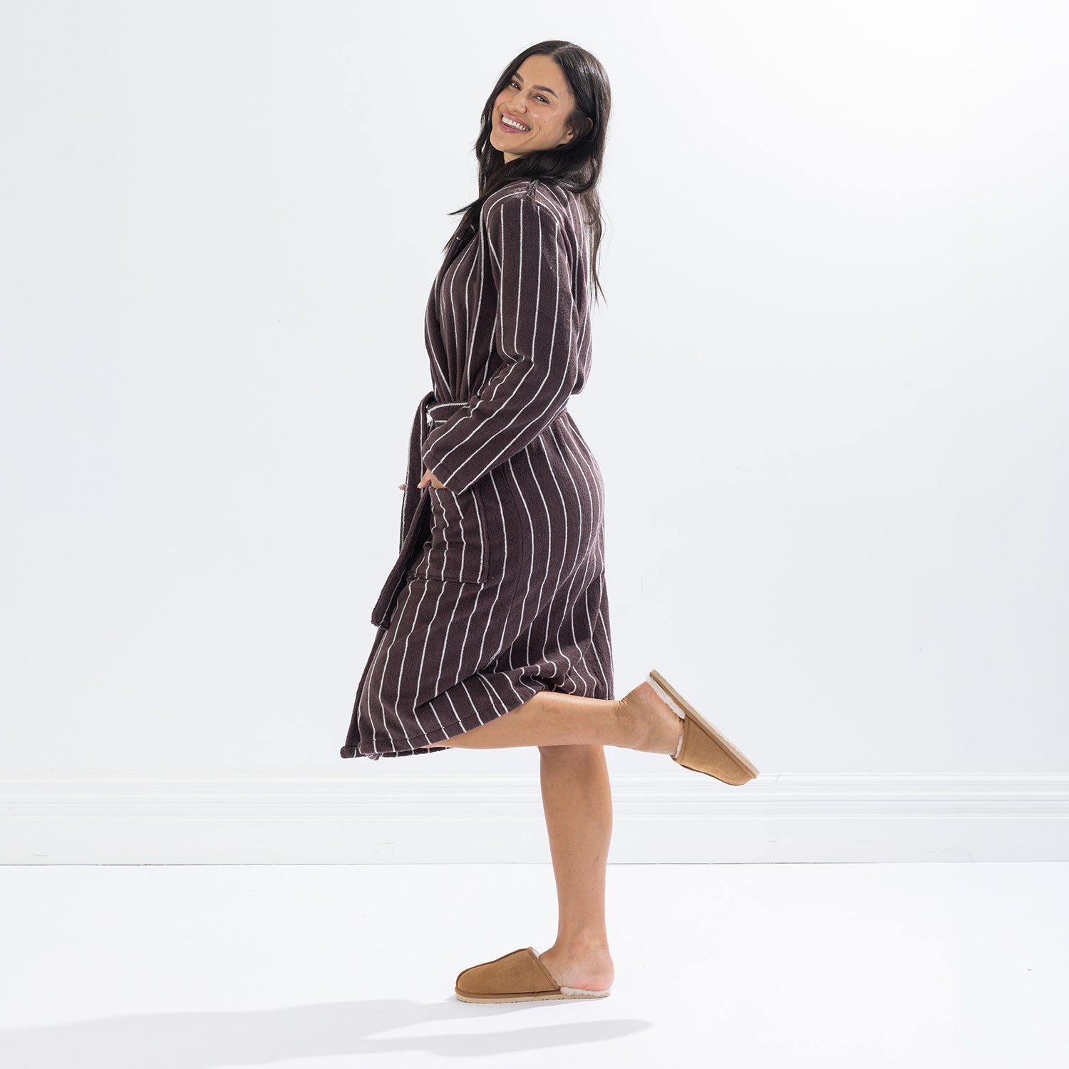Side profile of a woman wearing a chocolate brown striped towelling robe, smiling with one leg raised, showcasing the robe's fit and length.