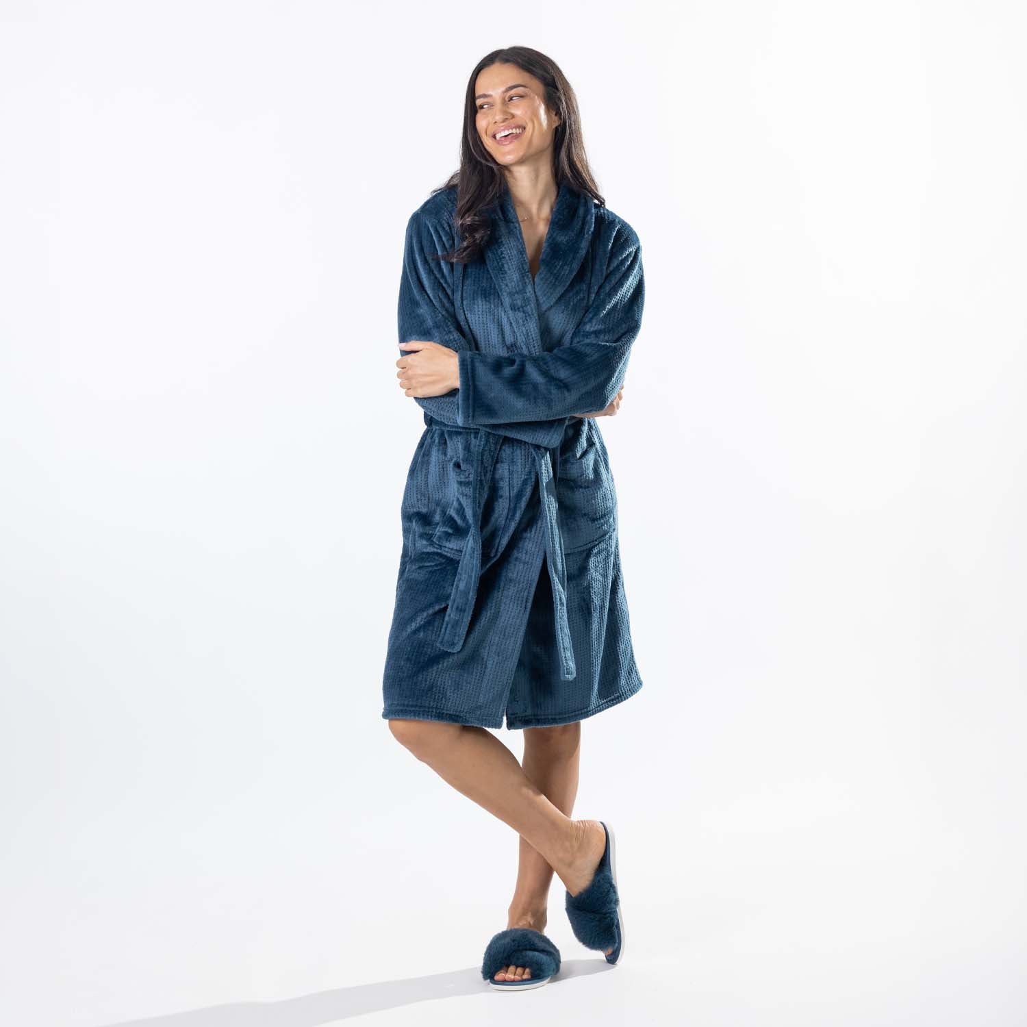 A woman wearing a plush teal waffle bathrobe with a shawl collar and tie belt, standing against a plain white studio background.