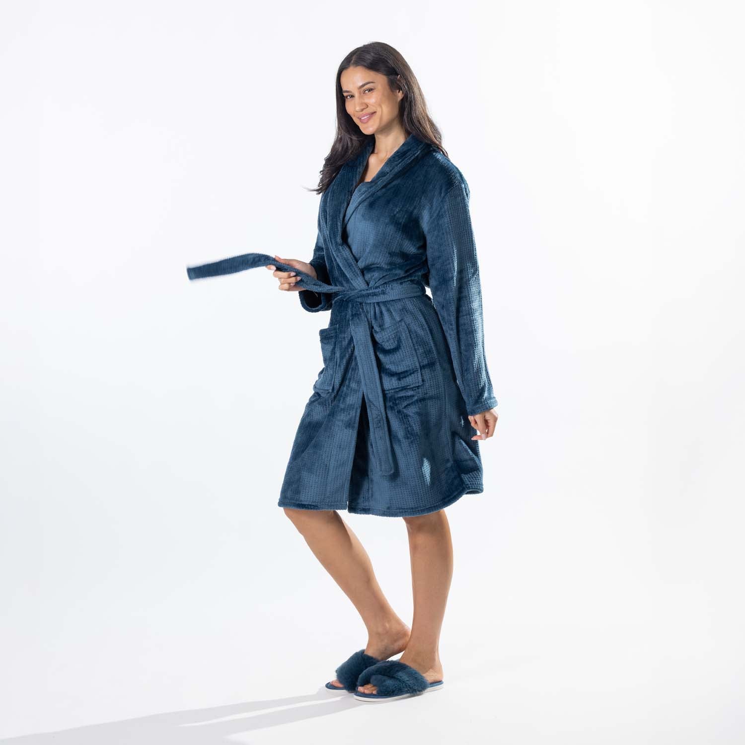 A woman wearing a plush teal waffle bathrobe with a shawl collar and tie belt, standing against a plain white studio background.