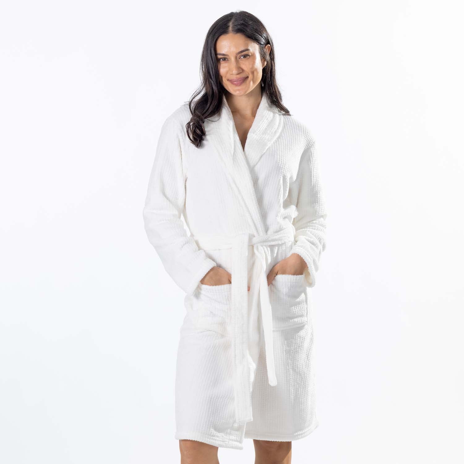 A woman wearing a white textured waffle bathrobe with a shawl collar, waist tie, and front patch pockets, standing against a plain white studio background.