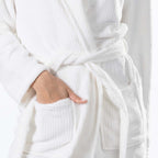 A close-up of a person wearing a white waffle-textured bathrobe, with one hand tucked into a front pocket.