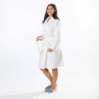 A woman modeling a white waffle bathrobe with a tie belt, standing against a clean white studio background and wearing blue slippers.