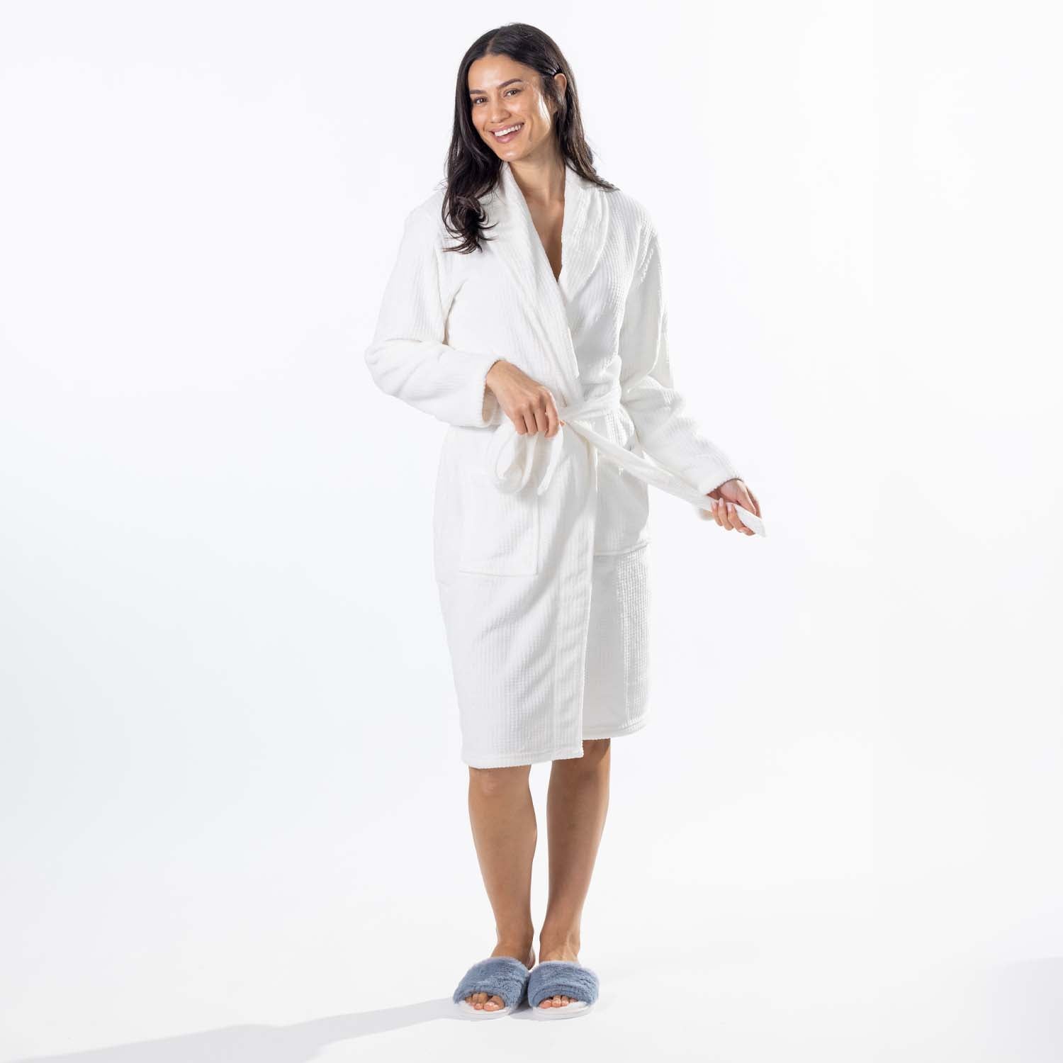 A woman modeling a white waffle bathrobe with a tie belt, standing against a clean white studio background and wearing blue slippers.