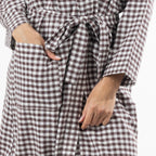 A close-up of a person wearing a brown and white gingham check robe with a tie waist and front pockets, with both hands visible in the pockets.