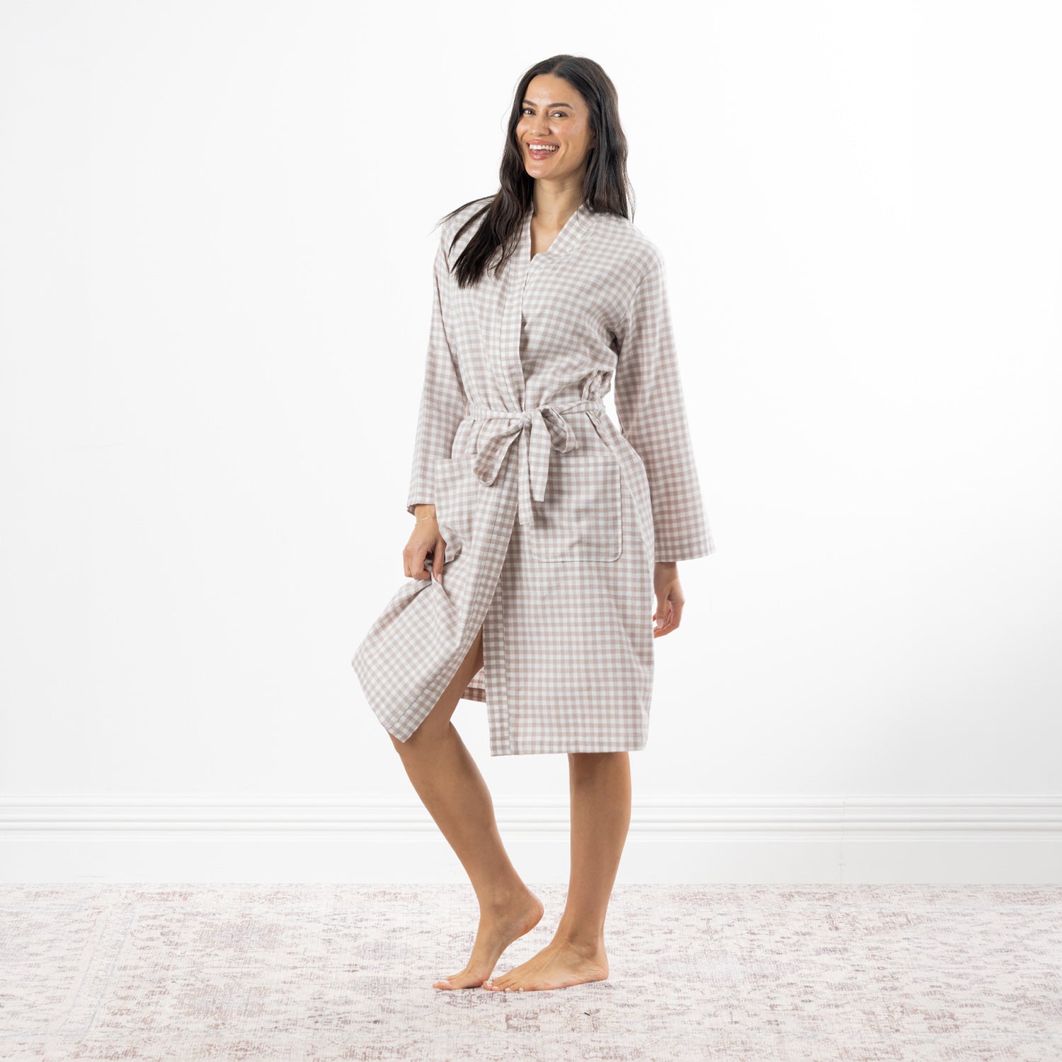 A woman modeling a natural and white gingham check bathrobe with a tie belt, standing barefoot on a light patterned rug against a white wall.