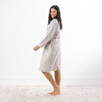 A woman modeling a long-sleeve, knee-length checkered robe with a tie waist, standing barefoot on a light patterned rug against a white wall.