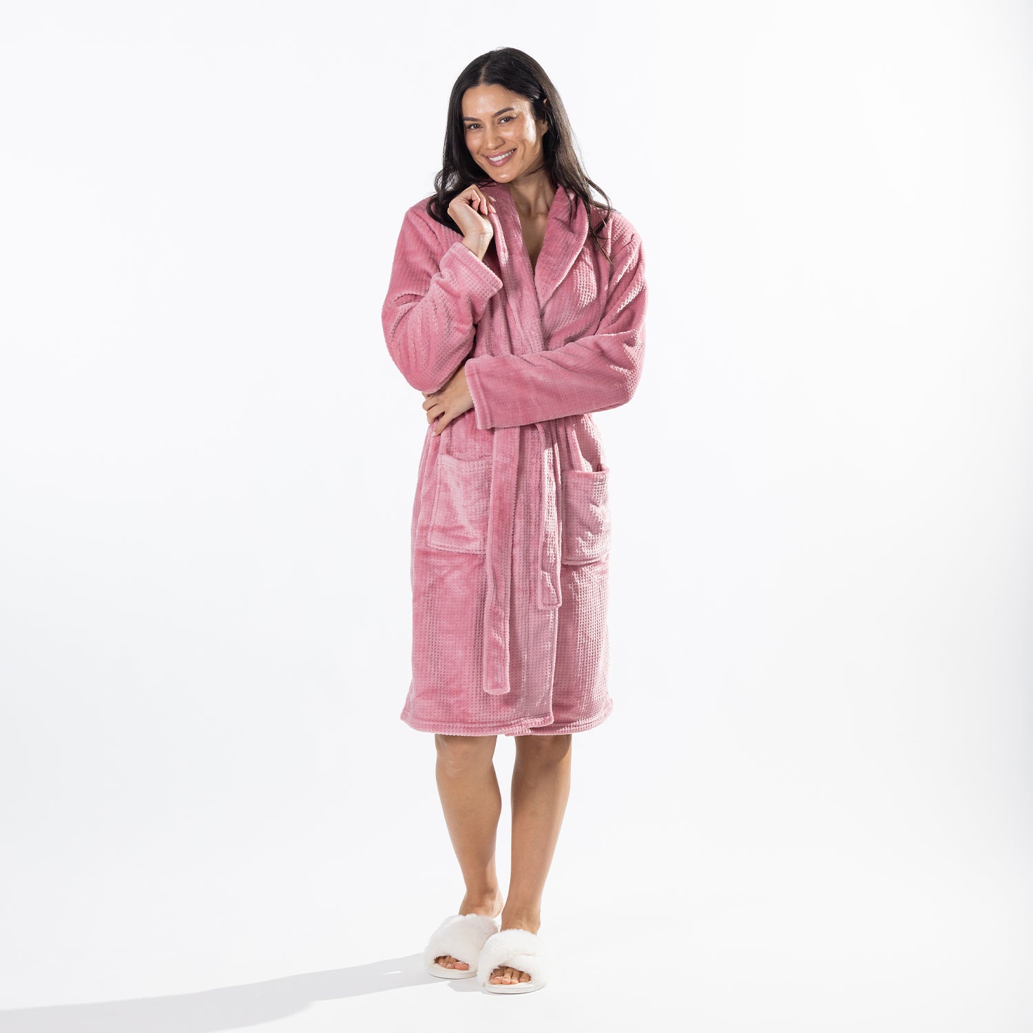 A woman wearing a plush pink bathrobe and white slippers, standing against a clean white studio background.