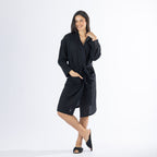 Full-length front view of a model wearing a black textured bathrobe with a wrap front, tie waist, long sleeves and front patch pockets, styled with black slippers on a white background.