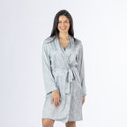 Amparo Cassie Satin Print Bathrobe in Denim modelled from the front, featuring a soft denim blue base with an all-over neutral and blue paisley print, long sleeves, a wrap front and matching waist tie.