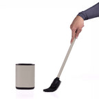 A hand holding a long-handled black and taupe cleaning brush next to a matching taupe and black cylindrical holder on a white background.
