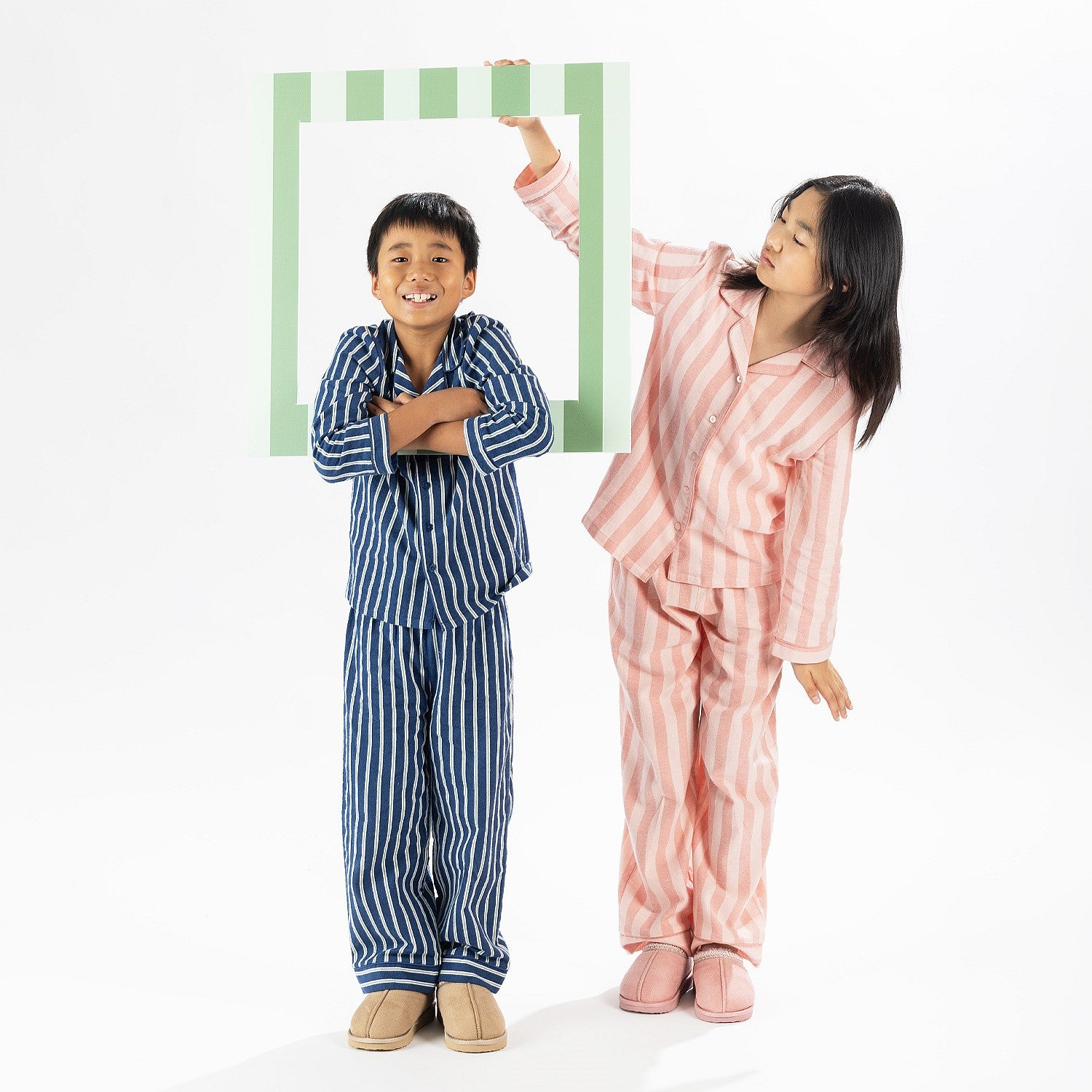 Two children wearing printed pyjama sets, one in pink stripes and one in navy stripe print, holding picture frame props.