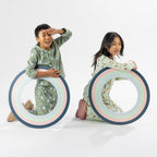 Two children wearing playful printed pyjama sets, one in green and one in cream, holding picture frame props.